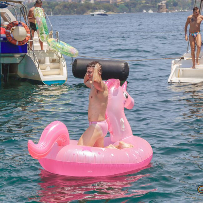 A person in swimwear rides a pink inflatable unicorn float in the water, holding a large black cylinder over their shoulder. Boats and people are visible in the background on a sunny day.