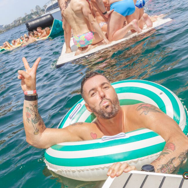 A man with tattoos poses in a green and white inflatable ring on the water, making a peace sign and puckering his lips. In the background, people are gathered on floating platforms, enjoying a sunny day.