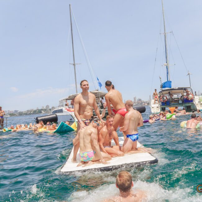 A group of men in swimsuits stand and sit on a floating mat in the water, surrounded by boats and people swimming and relaxing. The scene is festive and sunny, with a city skyline visible in the background.