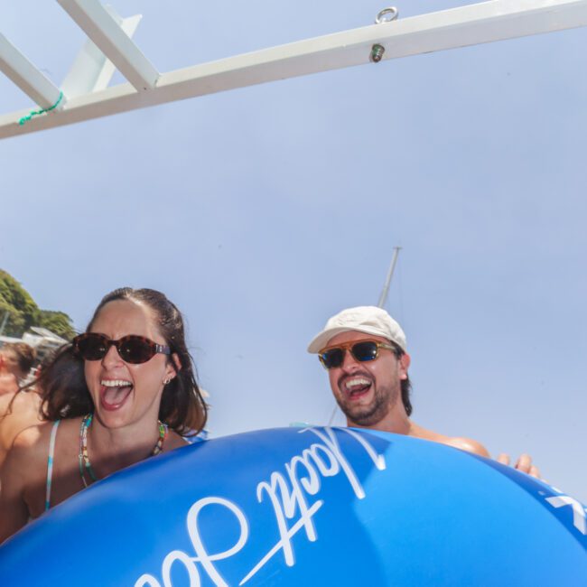 Two people wearing sunglasses laugh and smile while holding a large blue inflatable float. The sky is clear and sunny, and part of a boat structure is visible above them. Logos are seen at the bottom right of the image.