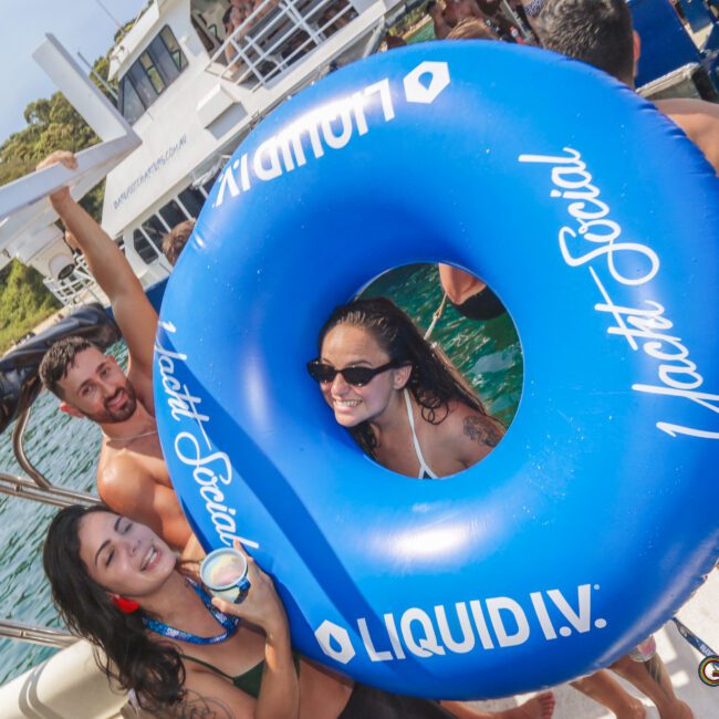 A group of smiling people are on a boat, enjoying a sunny day. One woman is inside a large blue inflatable swim ring that reads "Yacht Social" and "Liquid I.V." Boats and green water are visible in the background.