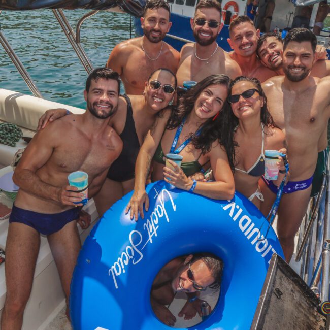 A group of smiling people in swimsuits pose together on a boat in the water, holding drinks and a blue inflatable tube. The sun is shining and everyone looks happy, enjoying a fun day out.