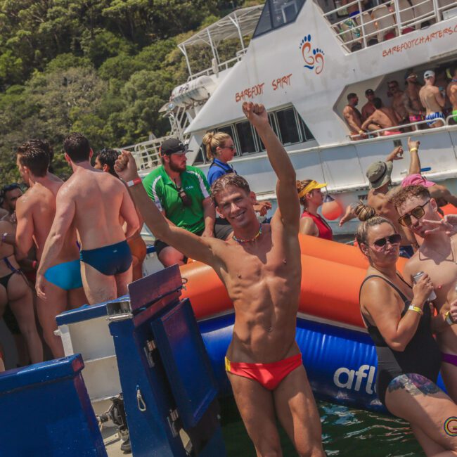 A crowd of people in swimsuits enjoy a sunny boat party on the water, with some dancing, smiling, and posing near an inflatable raft and a white boat in the background.
