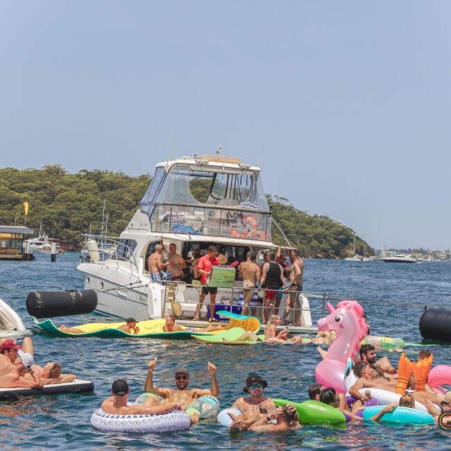 A lively group of people enjoy a boat party on a sunny day, relaxing on inflatables in the water, including a pink unicorn float, near several yachts with a forested shoreline in the background.