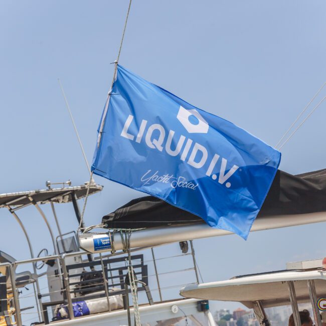 A blue flag with "LIQUID I.V." and "Yacht Social" printed on it flutters on a boat, with other boats and a clear sky visible in the background.