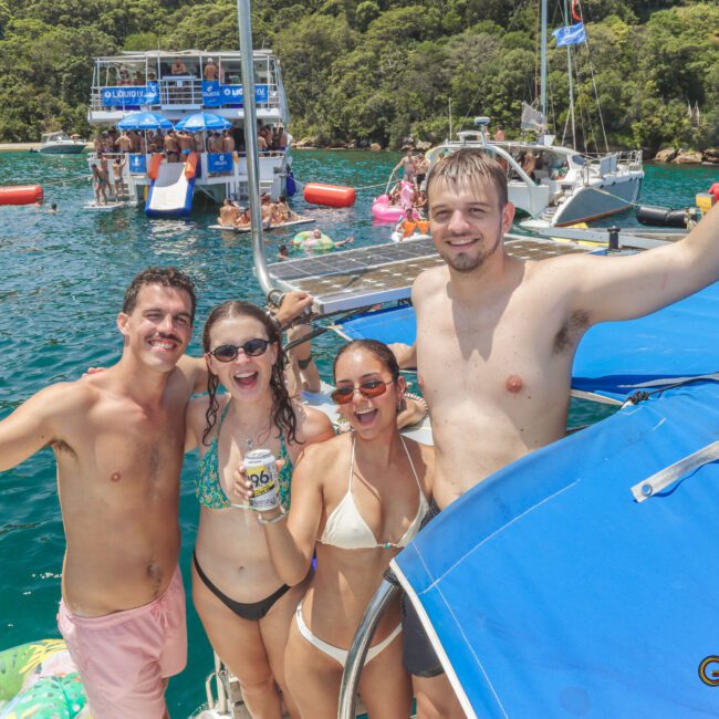 Four people in swimsuits smile and pose on a boat, holding drinks. Other boats and people are on the water behind them, with lush green trees in the background. It's a sunny day, and the atmosphere appears lively and festive.
