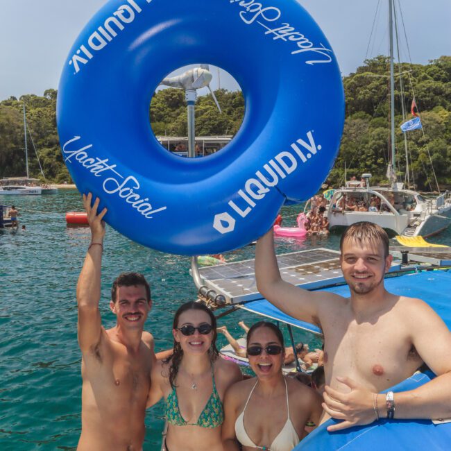 Four smiling young adults in swimsuits stand on a boat by clear water, holding a large blue inflatable ring that reads “LIQUID I.V.” Other boats and people are visible in the sunny background.