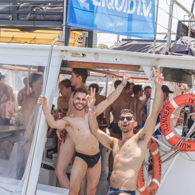 Two men in swimwear pose and smile on the deck of a boat, holding peace signs. Several people in swimwear are gathered inside the boat. A "LIQUID I.V." banner and life rings are visible in the scene.