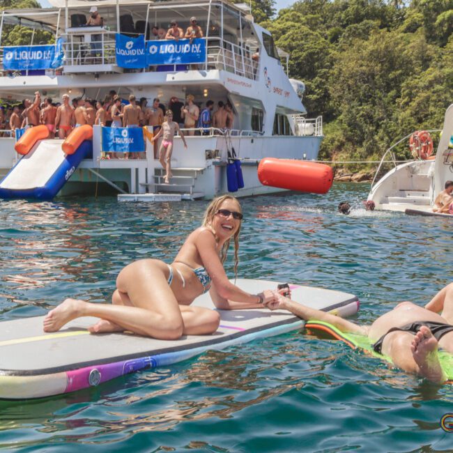 Two people relax on floating mats in the water near boats filled with people partying. A woman in a bikini smiles at the camera. In the background, others dance and socialize on the decks under sunny skies.