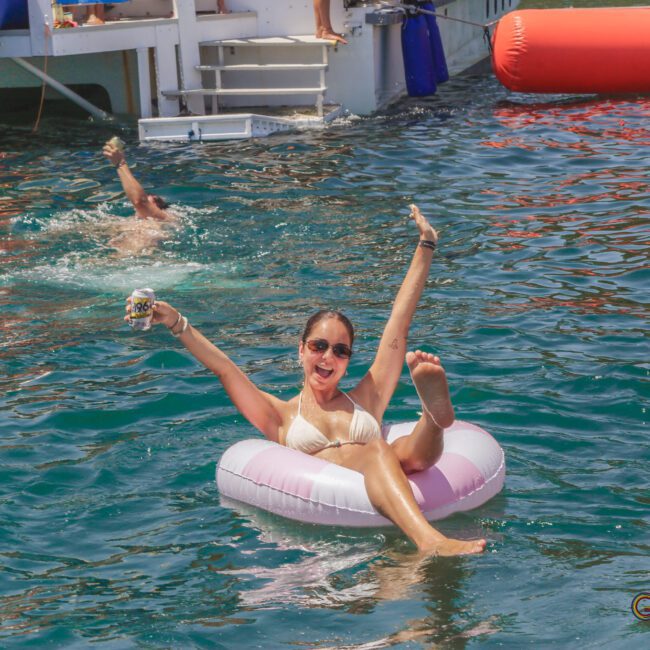A smiling woman in sunglasses and a white swimsuit lounges on a pink and white inflatable ring in the water, raising her arms. A person swims nearby, and a boat with inflatables is in the background.