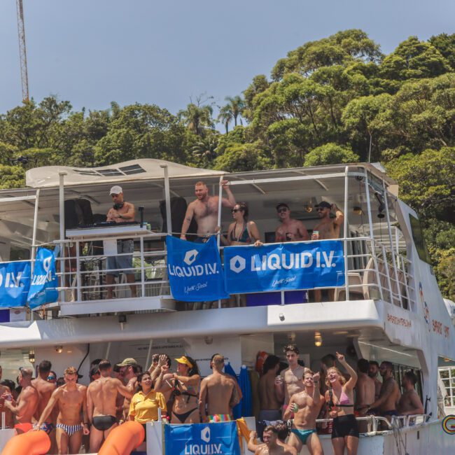 A group of people in swimwear are partying on a boat decorated with blue Liquid I.V. banners, surrounded by lush green trees and a sunny sky.