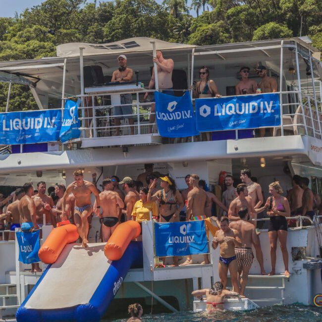 A large group of people in swimwear enjoy a lively party on a double-decker boat decorated with blue banners, anchored near a lush, green shoreline. Some are on the boat, while others swim or relax in the water.
