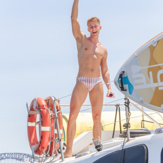 A smiling man wearing striped swim briefs stands on a boat, waving with one arm raised. There are life rings and a paddleboard beside him. The sky is clear and a bridge is visible in the background.
