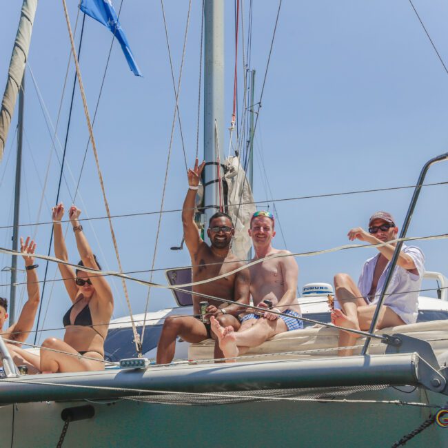A group of people in swimwear sit and smile on the deck of a sailboat, waving and enjoying the sunny weather under a clear blue sky.