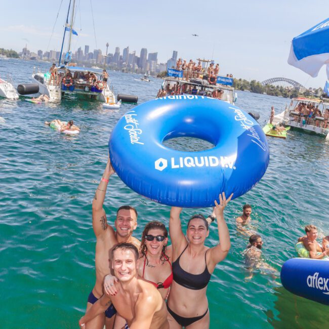 Four people in swimsuits smile and pose on a floating platform in the water, holding a large blue inflatable tube above their heads. Other swimmers and boats are visible in the background, with a city skyline further away.