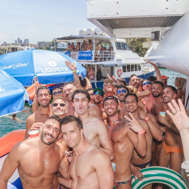 A large group of smiling men in swimwear gather for a photo at a lively pool or boat party, surrounded by floaties and blue "LIQUID I.V." umbrellas, with boats and water in the background on a sunny day.