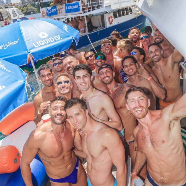 A large group of smiling men in swimwear pose together on a crowded boat deck during a sunny day, with inflatable pool toys and another boat visible in the background.