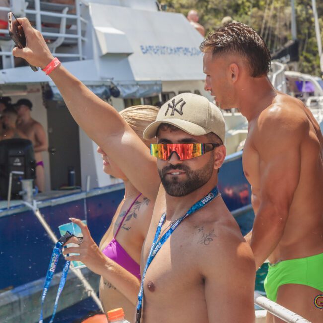 A shirtless man wearing sunglasses and a cap raises his phone for a selfie on a boat with other people in swimwear. In the background, more people are standing on another boat under sunny weather.
