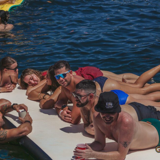 A group of people in swimsuits relax and smile while lying on a floating mat on the water, enjoying a sunny day. Some have drinks in hand, and others wear sunglasses. The atmosphere is lively and carefree.