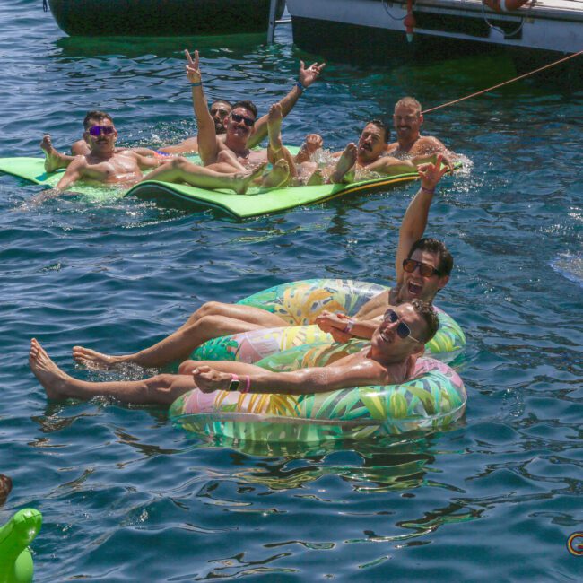 A group of people enjoy a sunny day on the water, relaxing on inflatable rafts and pool floats, smiling and making peace signs near several boats.