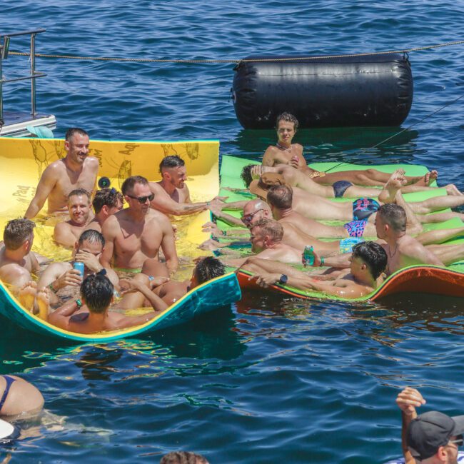 A group of men in swimsuits relax and socialize on floating mats in blue water under bright sunlight, with some holding drinks and a black inflatable barrier nearby.
