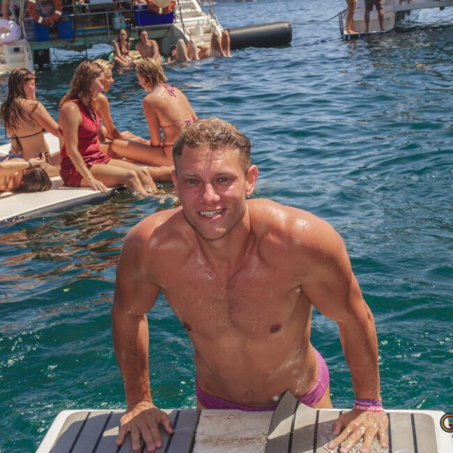 A smiling man in purple swim trunks climbs onto a floating dock from clear blue water, with other people relaxing and swimming nearby; boats are anchored in the background on a sunny day.