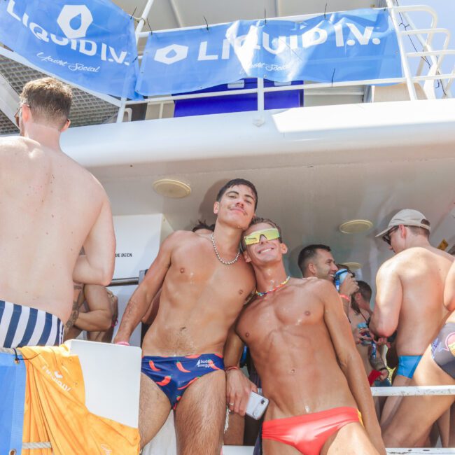 Two men in colorful swimwear and sunglasses pose and smile on a sunny boat deck with others around. Blue LIQUID I.V. banners and towels hang above and behind them, creating a festive summer atmosphere.