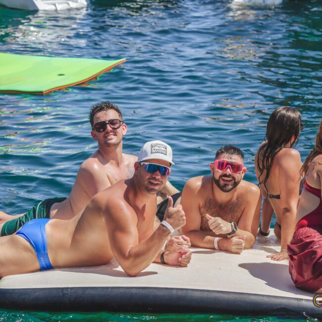 A group of young adults in swimsuits relax on a floating mat in a body of water, smiling and posing for the camera; some are giving a thumbs up while others sit and chat, enjoying a sunny day.