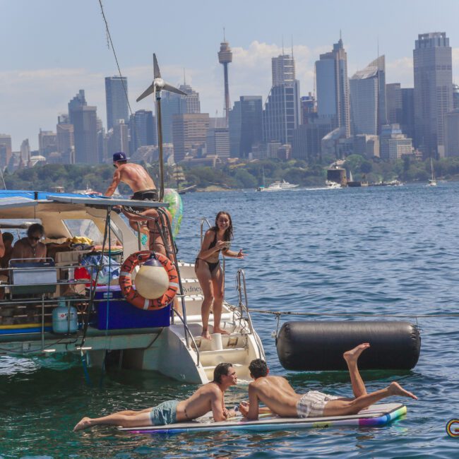 People relax and socialize on and around a catamaran in a harbor, with two people lying on a paddleboard in the water. The city skyline, including tall buildings, is visible in the background under a sunny sky.