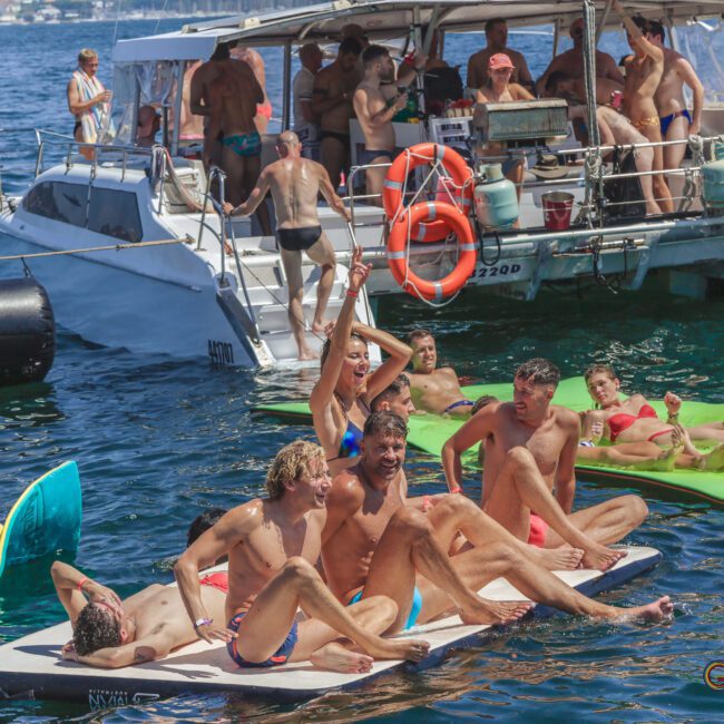 A group of people in swimsuits relax and have fun on floating mats in the water near a boat on a sunny day. Some people are sunbathing and others are socializing or swimming in the background.