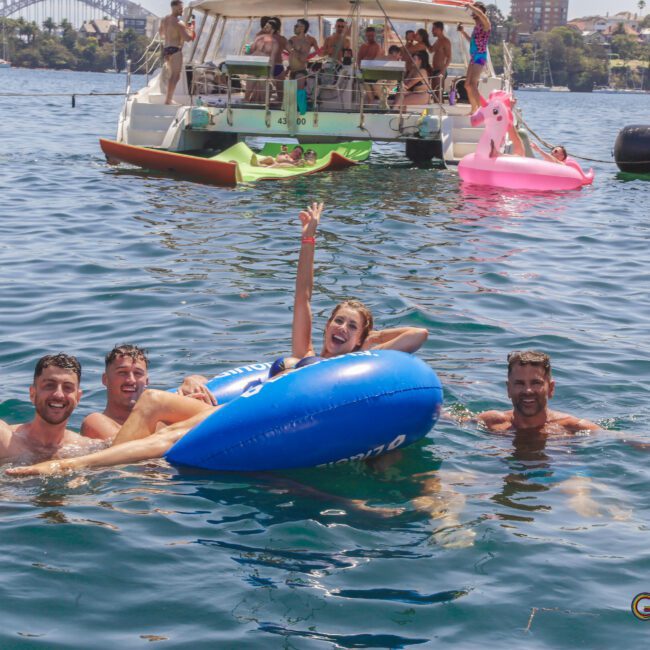 Four people smile and pose in the water around a blue inflatable float, while others relax on boats and pool floats in the background on a sunny day. The scene is festive and joyful.