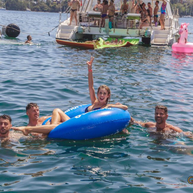 Four people smile and wave while relaxing on blue inflatables in a lake, with boats and other people enjoying the water and sun in the background.