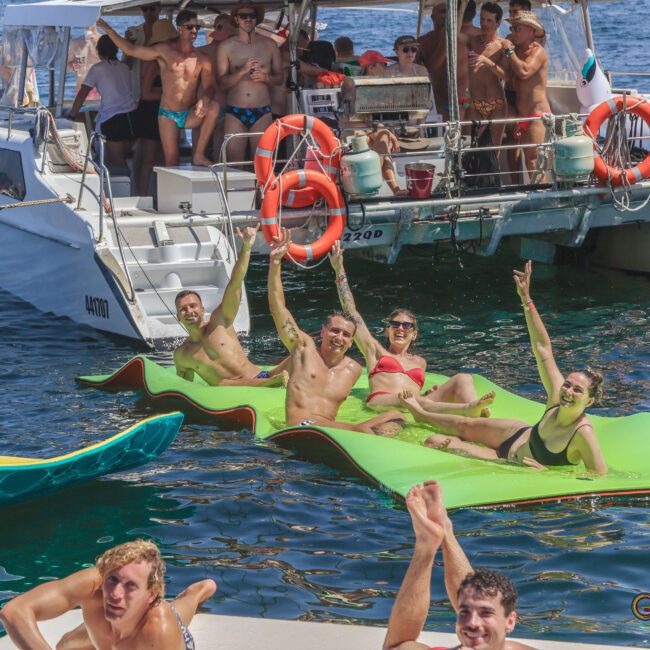 A group of people on a boat and lounging on float mats in the water, enjoying a sunny day. Some wave and smile at the camera, while others relax or swim, creating a lively and festive atmosphere.