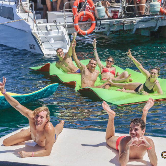 A group of smiling people in swimwear relax and pose on floating mats and a platform in the water near a docked boat, enjoying a sunny day. Some wave and flash peace signs while others make heart shapes with their hands.