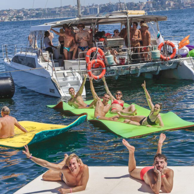 A group of people in swimsuits relax and pose cheerfully on floating mats near a boat on blue water, with more people aboard the boat. The sky is clear and a city skyline is visible in the background.