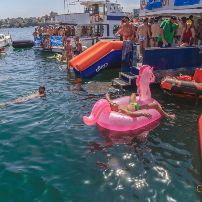 A person lounges on a pink inflatable unicorn in the water near boats filled with people in swimwear. Others swim nearby, and bright orange floats and rafts are visible. It’s a lively, sunny scene on the water.