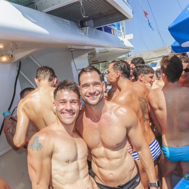 Two smiling shirtless men pose on a crowded boat party with other people in swimwear. Blue and white umbrellas and a sunny sky are visible in the background. The atmosphere is festive and lively.