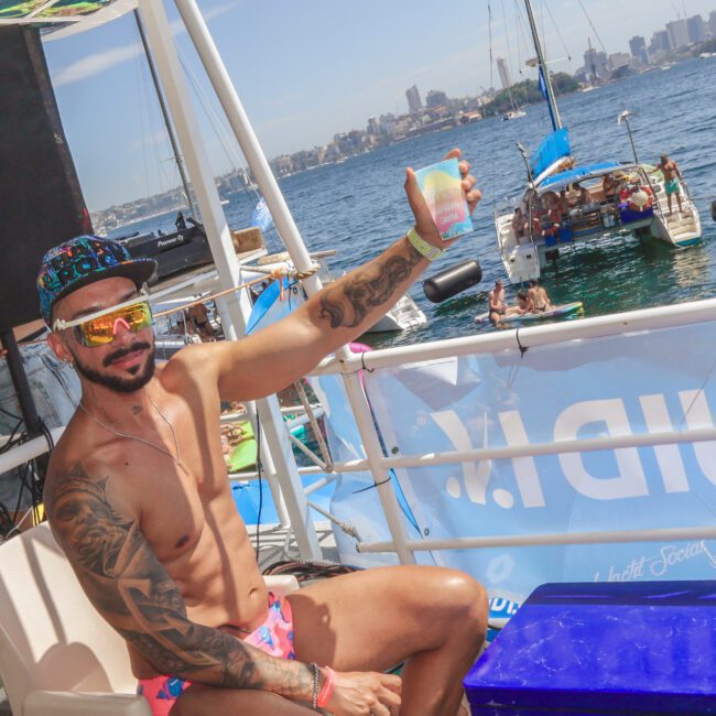 A man in swim trunks and sunglasses sits on a boat, smiling and holding up a drink. Other people relax on a nearby boat in the water, with a city skyline visible in the background under a sunny sky.