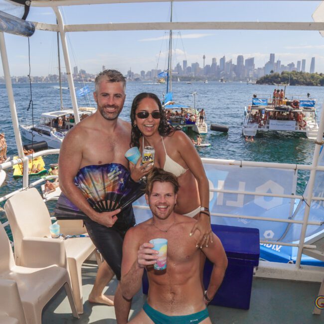 Three smiling people in swimwear pose on a boat with drinks, surrounded by other boats on the water. The Sydney city skyline is visible in the background on a sunny day.