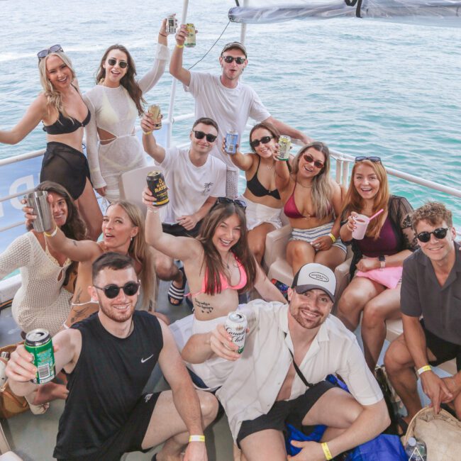 A group of smiling young adults on a boat, holding drinks and posing for the camera. They are wearing summer clothes and sunglasses, with water and a shoreline visible in the background.