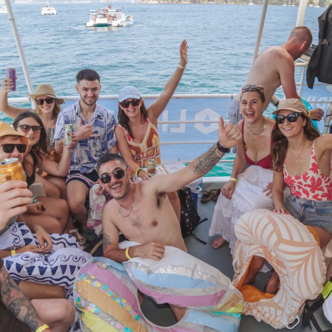 A group of smiling young adults in swimsuits and summer clothing relax and pose for a photo on a boat, raising drinks and waving, with the ocean and other boats visible in the background.