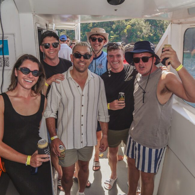 Six people in casual summer clothes and sunglasses smile and pose for a photo on a boat, holding drinks. There is water and greenery outside the boat window, and sunlight fills the scene.