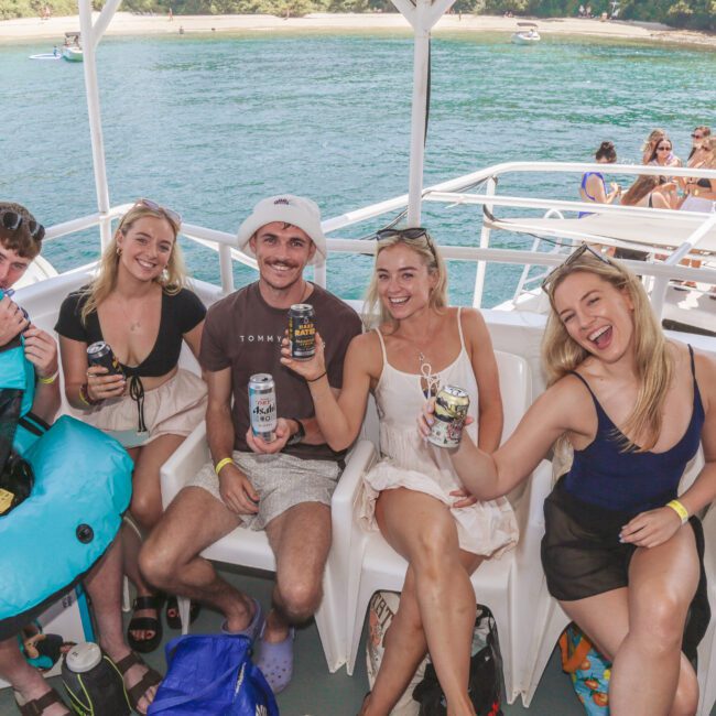 Five young adults sit on a boat, smiling and holding drinks. One person holds a blue inflatable tube. The boat is on clear blue water near a lush, green shoreline, and other people are visible in the background.