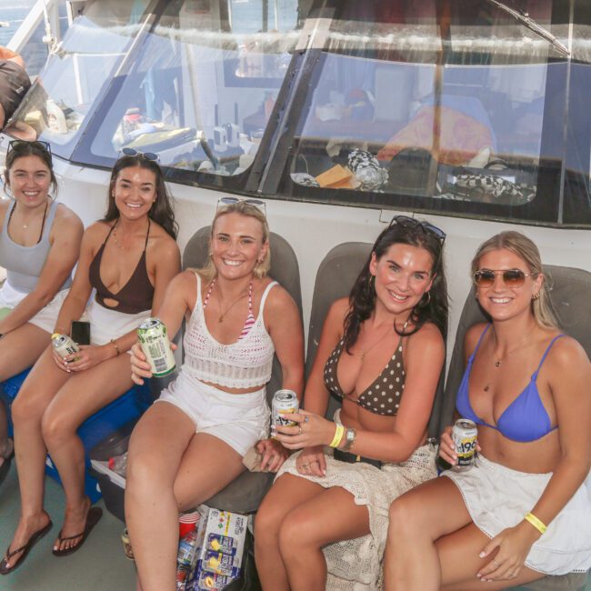 Five women in swimsuits sit and smile on a boat, each holding a can of drink. The sea and other people are visible in the background. It’s sunny, and the group appears to be enjoying a social outing.