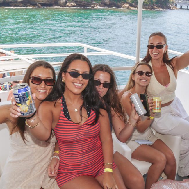 Five smiling women in swimsuits and sunglasses sit together on a boat, holding cans of drinks. Water and trees are visible in the background under a sunny sky.