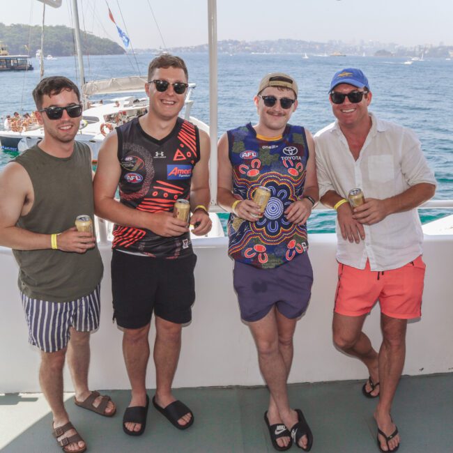 Four men in casual summer clothes and sunglasses stand on a boat deck, smiling and holding drinks. The background shows water, another boat, and a distant city skyline under a clear sky.
