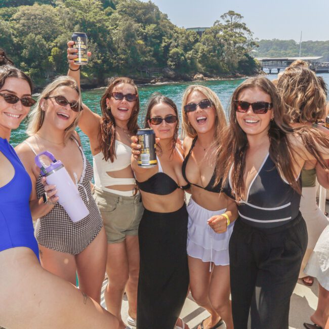 A group of six women in swimsuits and sunglasses smile and pose together on a sunny boat party, holding drinks, with blue water and lush trees in the background.