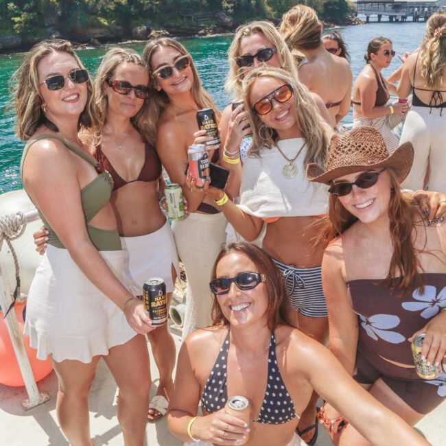 A group of women in swimsuits and sunglasses smile and pose together on a boat, holding drinks. Trees and water are visible in the sunny background, giving a festive summer atmosphere.