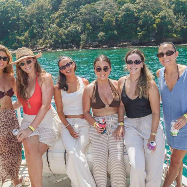 Six women in summer outfits sit on a boat railing, smiling at the camera, holding drinks. Behind them is clear turquoise water and a lush green forested shoreline under bright sunlight.