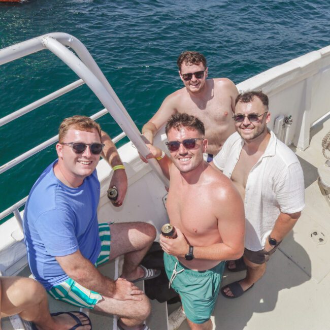 Four smiling men wearing sunglasses, summer clothing, and holding drinks pose together on the deck of a boat under bright sunlight, with blue ocean water visible in the background.